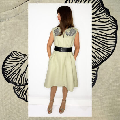Light green sleeveless dress with Djamoer Barat print, featuring a vegan black belt, side-seam pockets, and black bias tape finishing.