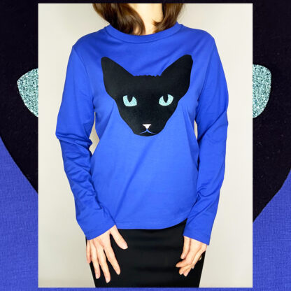 A long-sleeved dreamy blue t-shirt featuring a hand-printed black velvety Sphynx cat design with turquoise glitter eyes and a small pink nose. Made from organic cotton.